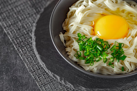 Udon noodles with raw egg and green onion in bowl, on slate stone plate round, dark background, selective focusの写真素材
