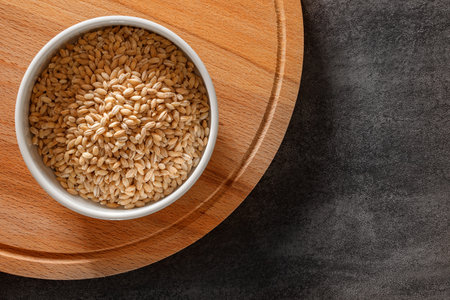 Pearl barley, raw grains in a white bowl on a wooden round stand, on dark background, top view, space to copy textの写真素材