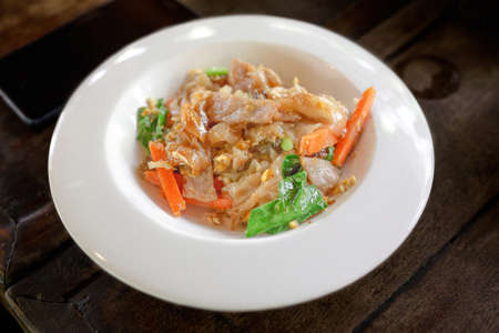 Stir fried pork noodles with black soy sauce in a white plate, old wooden table backgroundの写真素材