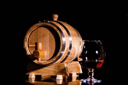 Glass of cognac with oak barrel on black background. Wooden barrel for whiskey with glass.の写真素材