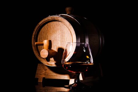 Glass of cognac with oak barrel on black background. Wooden barrel for whiskey with glass.の写真素材