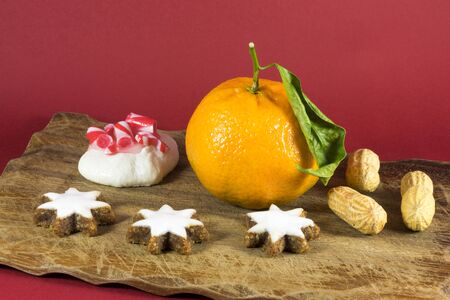 Seasonal Christmas still life home decoration with a fresh clementine, groundnuts or peanuts, white iced star cookies and a festive meringue with candy cane topping on a red backgroundの写真素材
