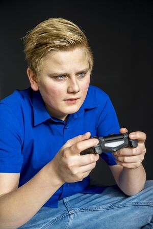 Serious young teen in blond hair, blue shirt and jeans sitting while holding video game controller...