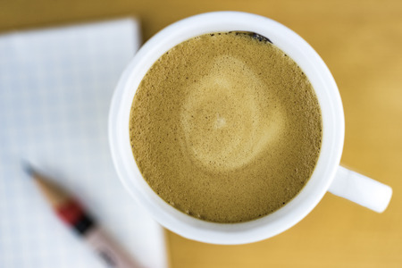 High Angle Still Life View of Cup of Frothy Coffee Resting on Wooden Table or Desk with Sharpened Pencil and Pad of Grid Paper Diffuse and Out of Focus in Backgroundの写真素材