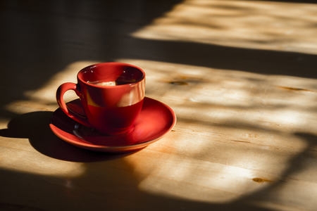 Profile Still Life of Red Cup of Coffee with Saucer on Wooden Kitchen Table with Bright Sunlight Streaming Through Window with Shadows and Ample Copy Spaceの写真素材