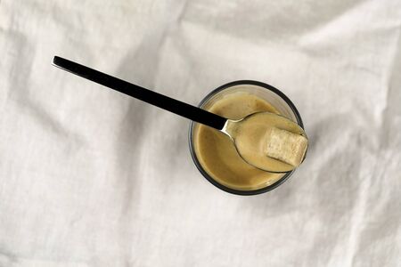 Glass mug of hot espresso coffee with a sugar cube suspended over the top in a teaspoon viewed from above on white fabric with copy spaceの写真素材