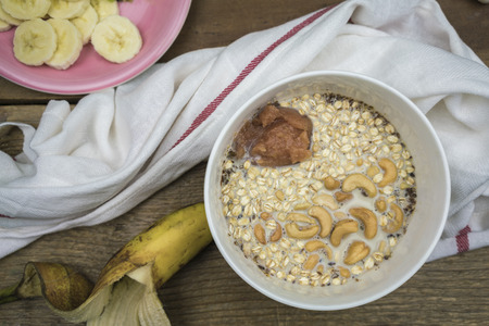 Oatmeal with milk, cashew nuts and applesauce in white ceramic bowl on wooden table with banana and white towel or napkin, healthy breakfast conceptの写真素材