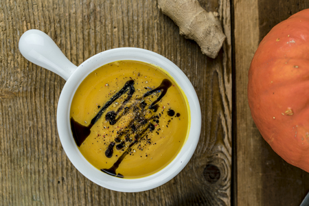 Seasoned pumpkin soup in ceramic white cup with pumpkin seed oil topping, viewed from above, with ginger on rustic wooden tableの写真素材