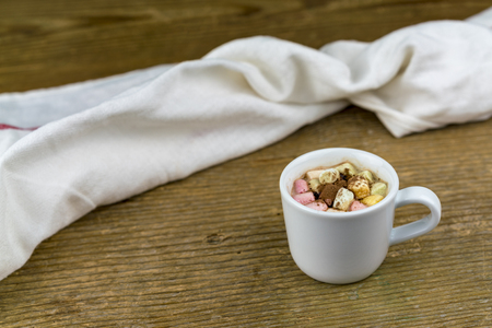 One white cup of cocoa with marshmallows of different colors, next to white fabric napkin nearby on old wooden table surface. Viewed on closeup with copy spaceの写真素材