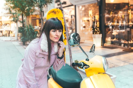 Portrait of a Stylish Pretty Young Woman in Autumn Fashion standing at the moped looking in the mirrorの写真素材
