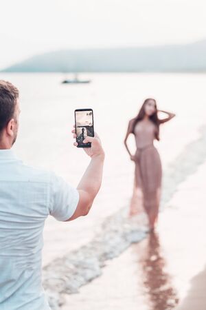 Man taking foto on a the camera on smartphone digital camera of a sporty young beautiful lady on a sea shore background. Outdoor lifestyle picture on a hot sunny summer day.の写真素材