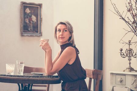 Toned picture of side view of beautiful blonde lady sitting in cafe or restaurant near window and drinking coffeeの写真素材