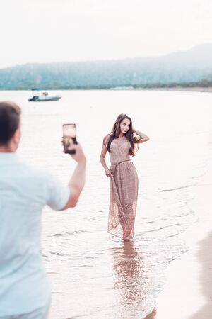 Man taking fotoon smartphone camera of a sporty young beautiful lady on a sea shore background. Outdoor lifestyle picture on a hot sunny summer day.の写真素材