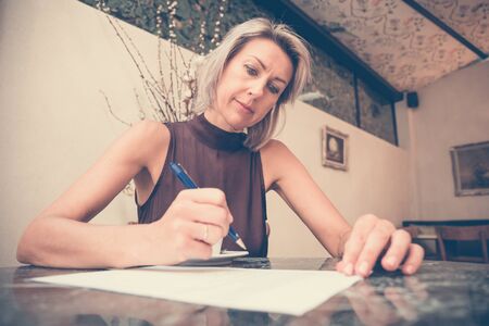 Toned picture of beautiful blonde lady sitting in cafe or restaurant near window and signs documentsの写真素材