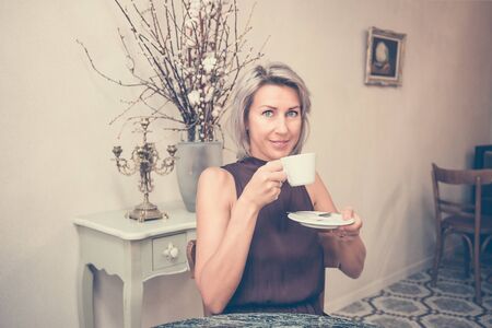 Toned picture of front view of beautiful blonde lady sitting in cafe or restaurant near window and drinking coffeeの写真素材