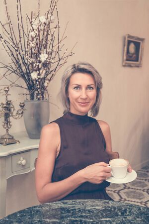 Toned picture of front view of beautiful blonde lady sitting in cafe or restaurant near window and drinking coffeeの写真素材