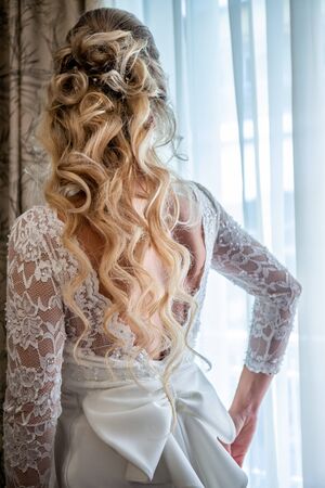 Beautiful bride in white wedding dress standing in her bedroom and looking in windowの写真素材