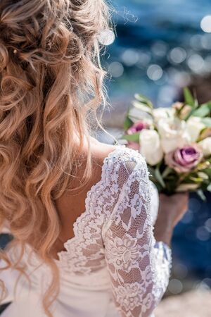 Beautiful bride in luxury wedding dress with bouquet at the sea side. Wedding by the sea. Bride walking around the sea near the place of the wedding ceremony. Back viewの写真素材
