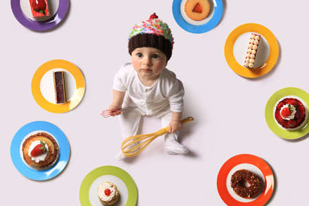 A baby holding two whisks and wearing a cupcake hat among plates of cakes.