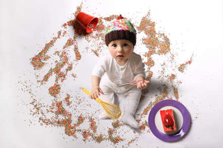 A baby holding two whisks and wearing a cupcake hat among sprinkles and a piece of cake.の写真素材