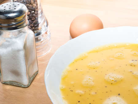 A plate with a beaten egg, a salt shaker, a pepper mill and an egg for omelette preparation