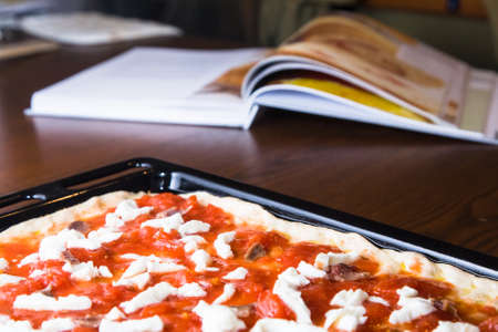 A raw pizza with mozzarella cheese, tomatoes and tuna ready to be put into the oven with recipe book in the backgroundの写真素材