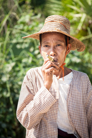 Inwa, Myanmar - 2016, December 25 : A Burmese middle-aged woman with a straw hat on smoking a green...