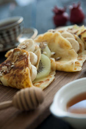 Crepe Waffle with Banana and Kiwi Wrapped on a Wooden Cutting Board with a Cup of Tea and Honey