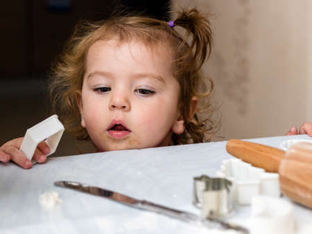 Baby girl is standing next to a kitchen table and looks at how the biscuit dough is doneの写真素材
