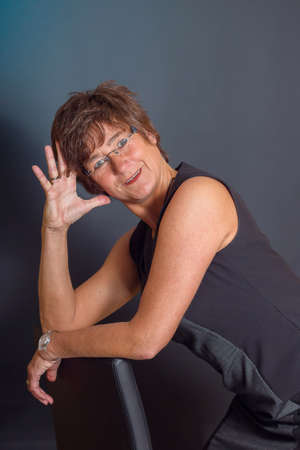 Smiling mature female with hand on her head seating opposite dark backgroundの写真素材