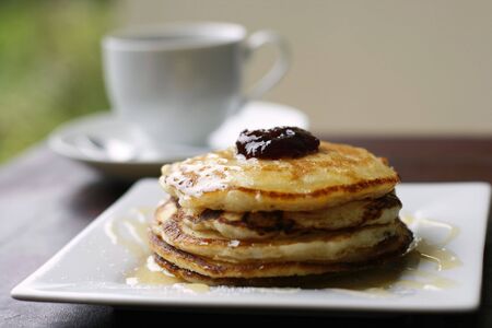 A stack of delicious golden pancakes with jam and honey on a white plate with a white coffee cup in the background.の写真素材