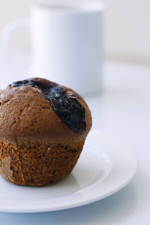 A delicious blueberry muffin on a white plate with a white coffee mug in the background.の写真素材