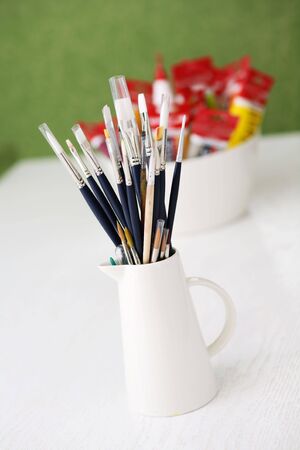 A set of paintbrushes in a jug and tubes of paint in a bowl on a white table in an outdoor setting with green grass in the background.の写真素材