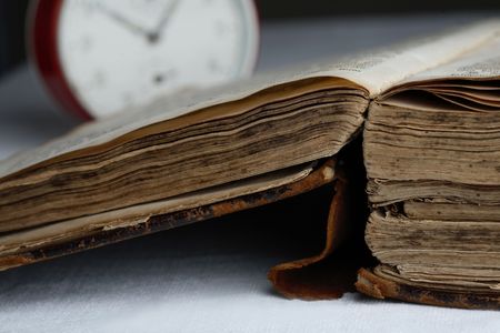 A close-up of an old, worn and damaged book with a dark brown hard leather cover and an old analog alarm clock in the background.の写真素材
