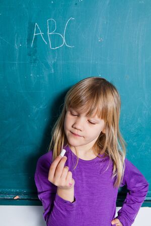 Young girl standing in front of chalkboard with letters of alphabet and looking at hand holding stick of chalkの写真素材