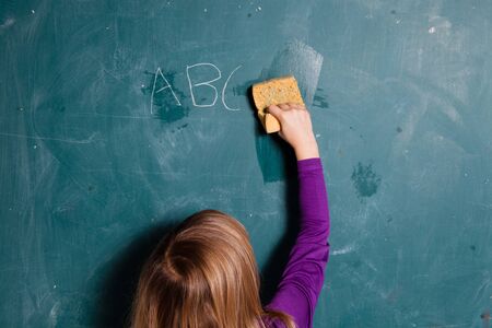 Young girl wiping letters written in chalk from chalkboard with wet spongeの写真素材