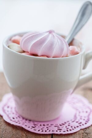 Closeup of cup of hot chocolate with pink meringue and spoon resting on small pink table mat on wooden tableの写真素材