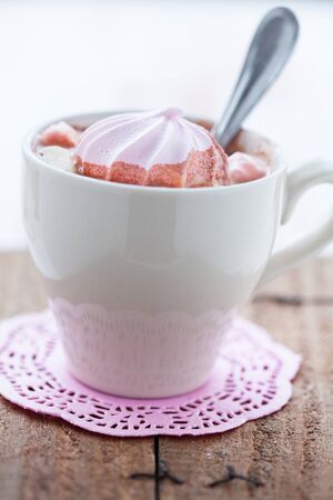 Closeup of cup of hot chocolate with pink meringue and spoon resting on small pink table mat on wooden tableの写真素材