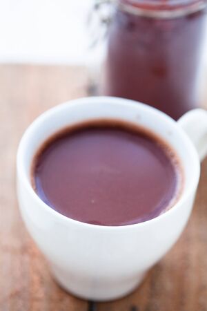 Closeup of cup of hot chocolate on a wooden table with cocoa powder in glass jar in backgroundの写真素材