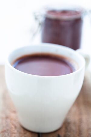 Closeup of cup of hot chocolate on a wooden table with cocoa powder in glass jar in backgroundの写真素材