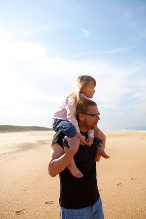 Father carrying young daughter piggyback on shoulders at beachの写真素材