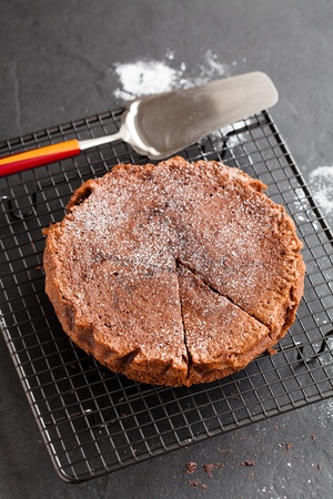 Freshly baked homemade chocolate cake on metal cooling rack with cut slice and cake server utensil on dark backgroundの写真素材