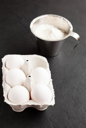 Chicken eggs in white carton with flour in metal measuring cup on dark backgroundの写真素材