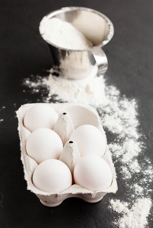 Chicken eggs in white carton with flour in metal measuring cup and spread over dark backgroundの写真素材