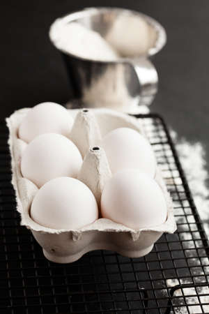 Chicken eggs in white carton on mesh tray with flour in metal measuring cup on dark backgroundの写真素材
