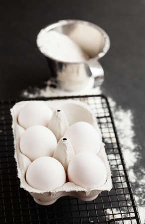 Chicken eggs in white carton on mesh tray with flour in metal measuring cup on dark background