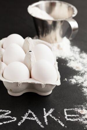 Chicken eggs in white carton with flour in metal measuring cup and spread on dark background with word bake written in chalkの写真素材