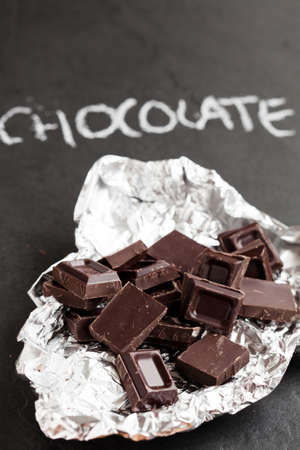 Closeup of chocolate squares in silver foil with word chocolate written in chalk on dark backgroundの写真素材