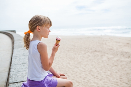 Young cute girl holding ice cream while sitting on wall at beachの写真素材