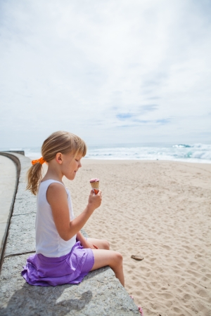 Young cute girl holding ice cream while sitting on wall at beachの写真素材